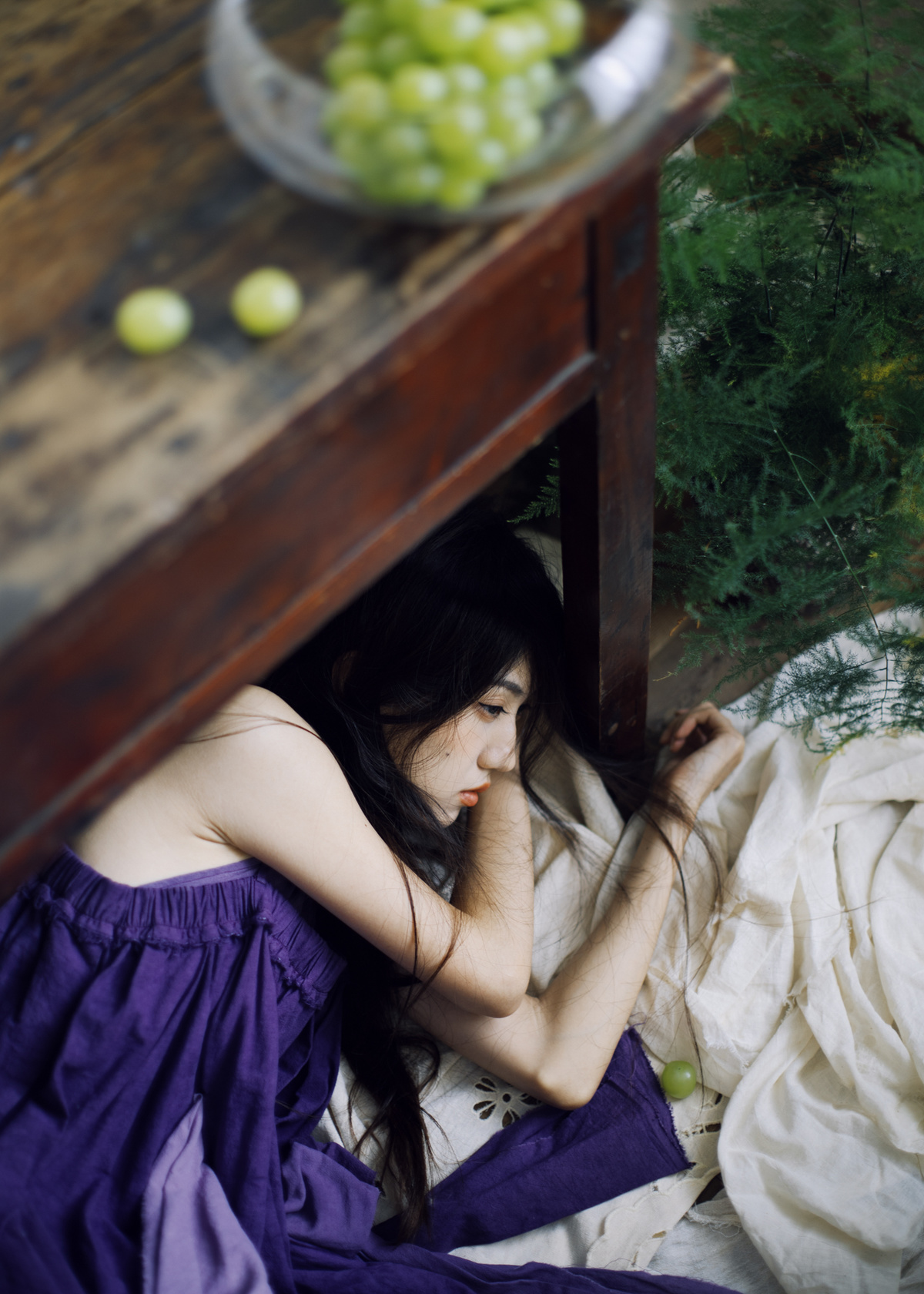 一个穿着紫色裙子的年轻女子躺在床上 盖着毛毯 桌子上放着葡萄和苹果。