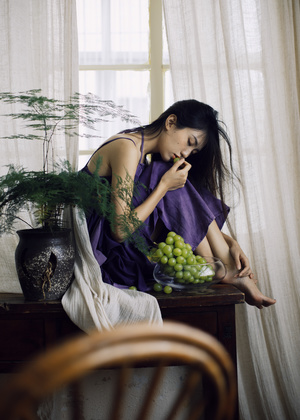 一个穿着紫色裙子的年轻女子坐在窗前的一把椅子上 桌子上放着一碗绿葡萄。