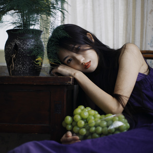 一名穿着紫色的年轻女子躺在床边 旁边放着一瓶绿色的葡萄。
