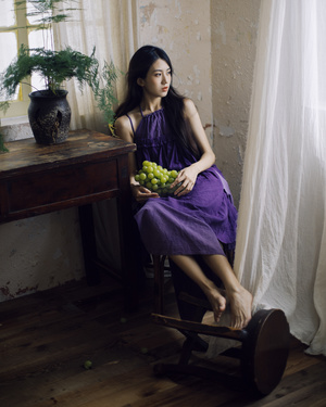 一个穿着紫色裙子的年轻女子坐在一个木椅上 在一个有窗户和木桌上有水果碗的房间里。