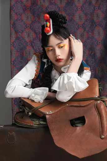 一位穿着复古裙子的长发女子 抱着棕色包包 坐在棕色箱子上。