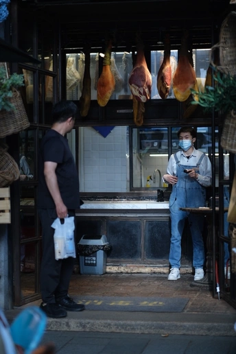 一个人戴着面具站在一个有火腿的商店前 一个顾客在看着。