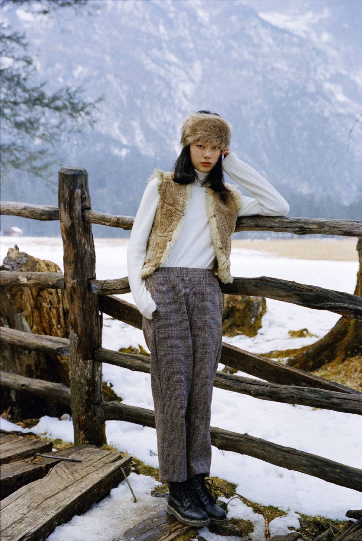 一位戴帽子和裤子的女人站在雪中的木质围栏前