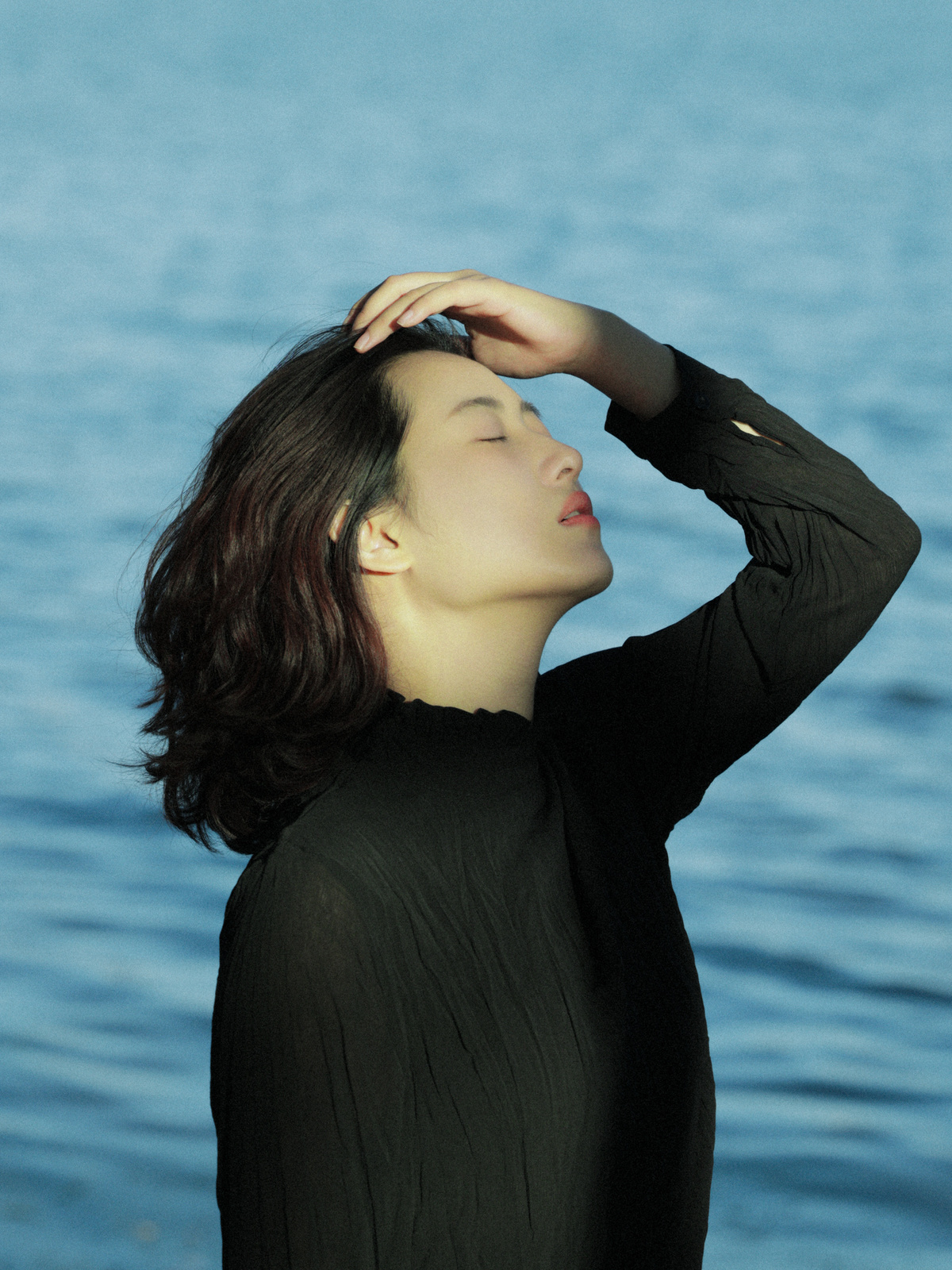 一位穿着全黑衣服的年轻女子闭着眼睛 头发在空中飘动 站在一个水域旁。