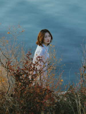 一位穿着白色衣服的年轻女子站在灌木丛和水体旁边 背景是天空。