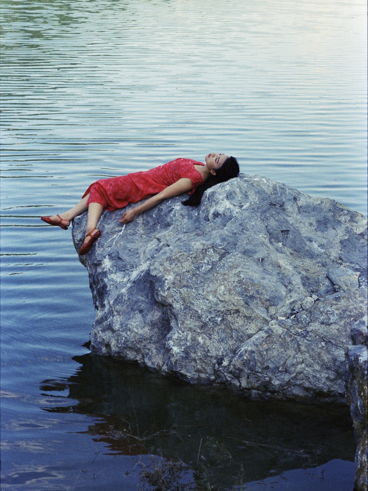 一位穿着红衬衫的年轻女子躺在一片水域中的一块大岩石上。
