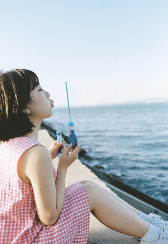一个穿着连衣裙吹泡泡的年轻女子坐在码头上眺望大海