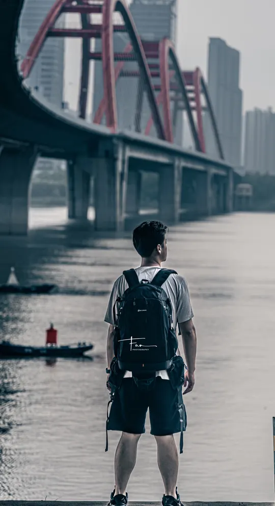 一个人背着书包站在一个有水体和桥作为背景的地方