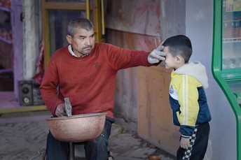 一个男子把一个碗举在年轻男孩的头上 这个男孩正在由一个小孩子理发。
