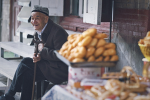 一个老人坐在桌子前的长椅上 手里拿着一根拐杖 桌子上放着食物。