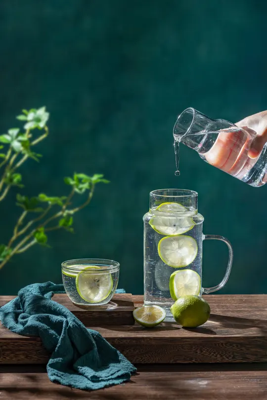 一只手从水桶中倒水入杯 杯中冰块、柠檬和青柠在木桌上 