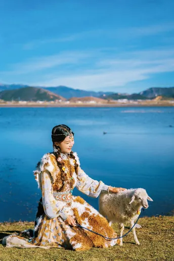 一个穿着裙子的年轻女孩坐在草地上 前面有狗和羊 在水边。