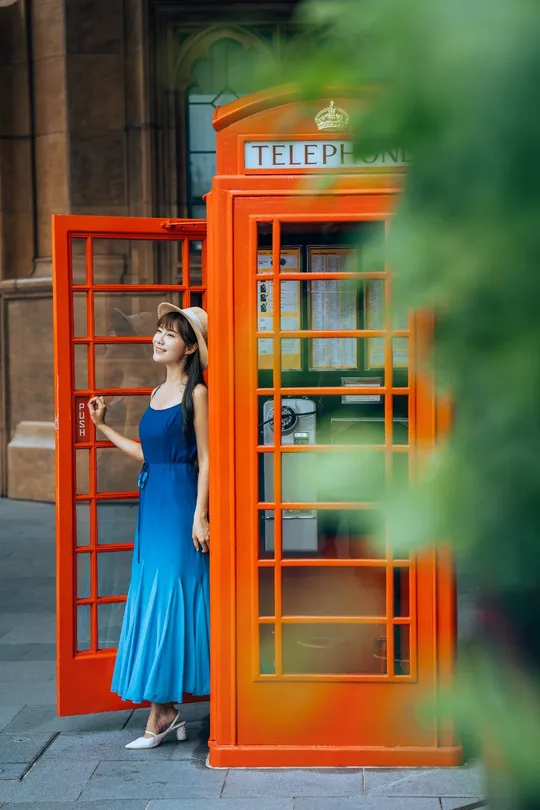 一位穿着蓝色连衣裙的年轻女子站在红色电话亭旁边