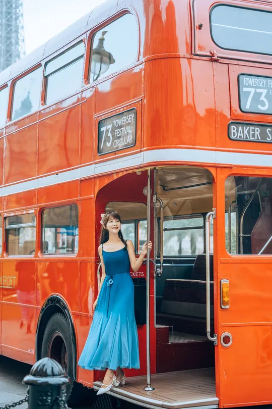一位穿着蓝色连衣裙的女性站在一辆红色的双层公交车旁边