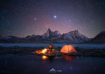一个人坐在湖边篝火旁 周围星空璀璨 远处山脉如画。