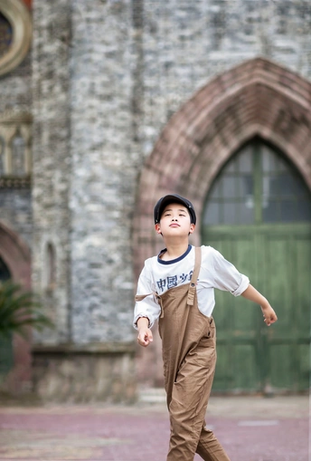 一个戴帽子的年轻男孩走在教堂前面的人行道上 手里拿着一个小滑板。