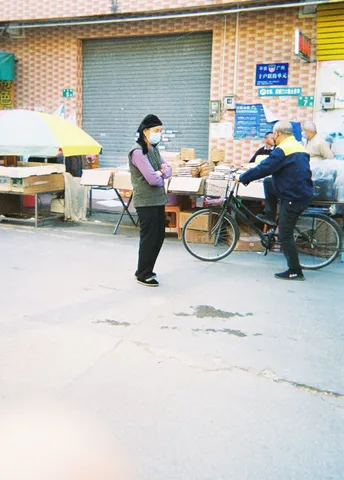 一名戴面具的女人走过一个市场 那里一个男人正在骑自行车把他的自行车推下街道。