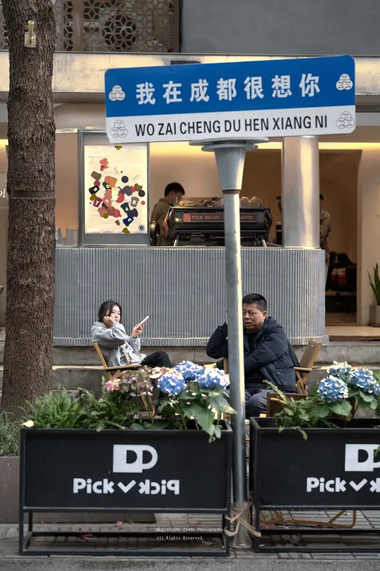 坐在餐厅前的长椅上 人行道上有一栋建筑 上面有标志 人行道上还有花。