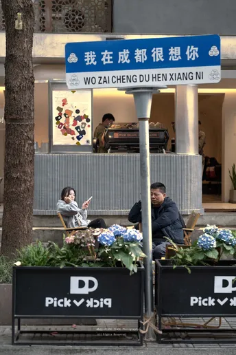 坐在餐厅前的长椅上 人行道上有一栋建筑 上面有标志 人行道上还有花。