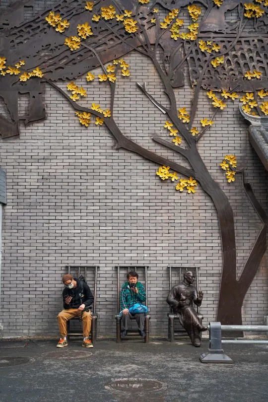 一些男人坐在建筑物前的一张长椅上 墙上有一棵树的雕像。