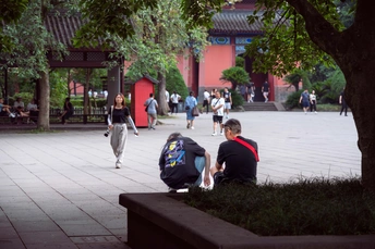 一个背着书包的年轻男孩坐在庭院的长椅上 背景有一些人在经过一座建筑。