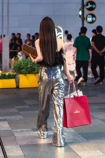 一位穿着银色裤子的年轻女子 提着红色包包走在街上。