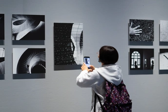 一个小女孩正在看手机上的照片 而一个妇女站在一幅挂满照片的墙前。