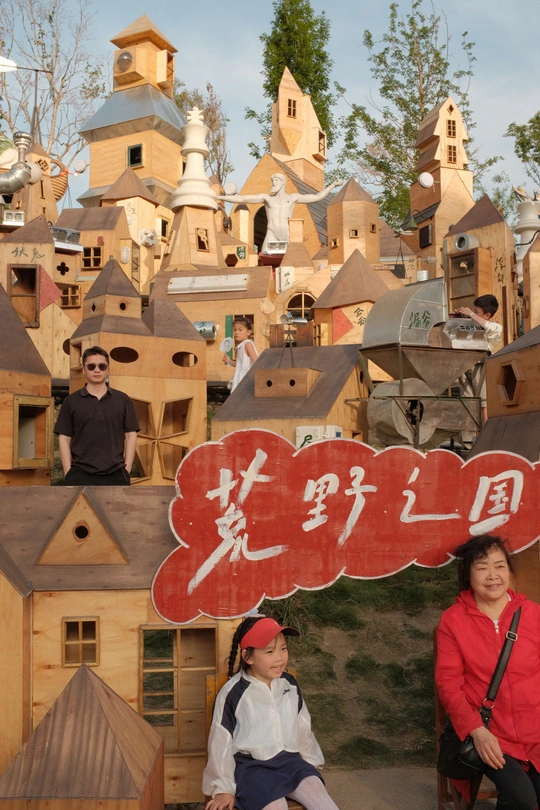 一位妇女和儿童在木质建筑群前为一张照片摆姿势 前面有一个写着hello的牌子 一群人在那里。