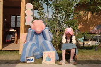 一个小女孩坐在蓝色房子的前面长凳上 一个粉红色的小玩具坐在粉红色玩具大象的前面一把椅子上。