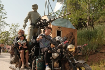 一个男子、一个女子和一个孩子骑在一辆摩托车上 在他们身后有一尊男子和女子的雕像。