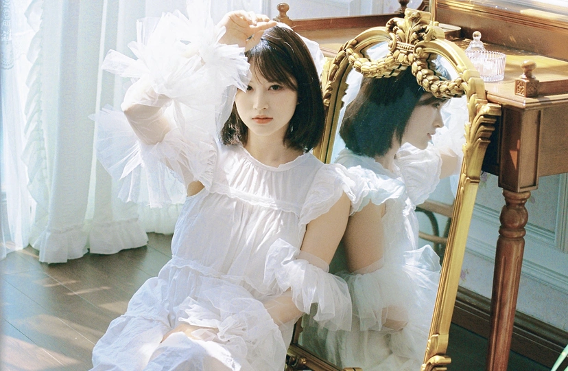 一位穿着白色连衣裙的年轻女子坐在镜子前的一把椅子上