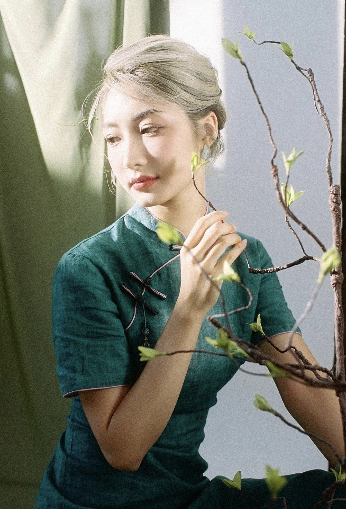 一位穿着绿色连衣裙的年轻女子正在靠窗姿势摆拍 同时看着植物的枝条 为画像做姿势。