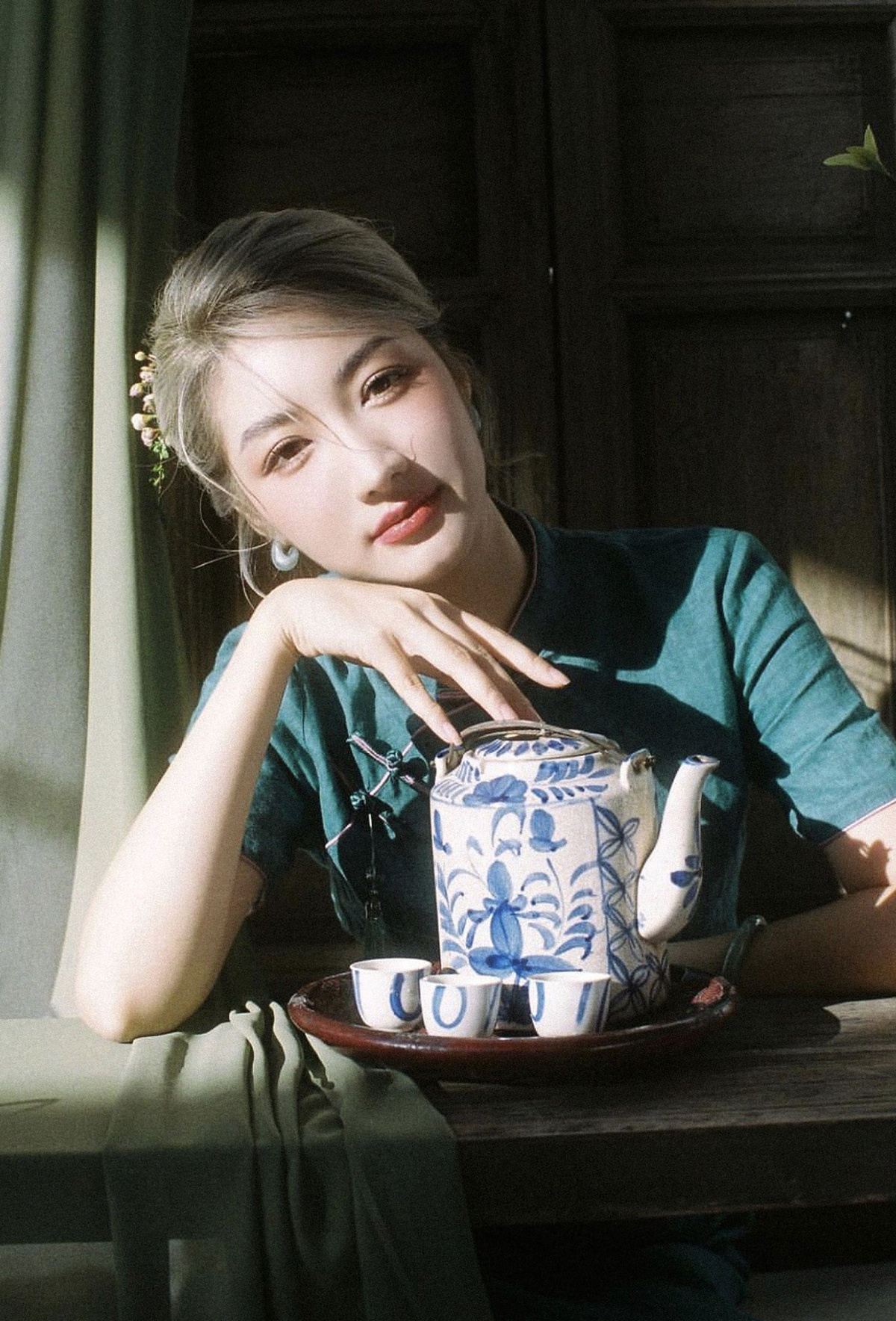 一个年轻女子坐在窗前一张桌子旁 手里拿着一对蓝色的杯子与托盘 里面盛着饮料。