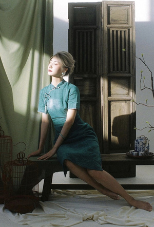 一个穿着绿色连衣裙的年轻女子坐在有窗户的房间里的一张长椅上 一只鸟儿正坐在地板上。
