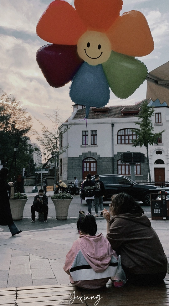 一位妇女坐在长凳上 小女孩正在注视着一只形状像花的气球 其他人在一旁观看。