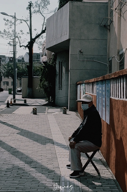 一个戴帽子的老人坐在人行道旁的一把椅子上 旁边有一栋建筑。
