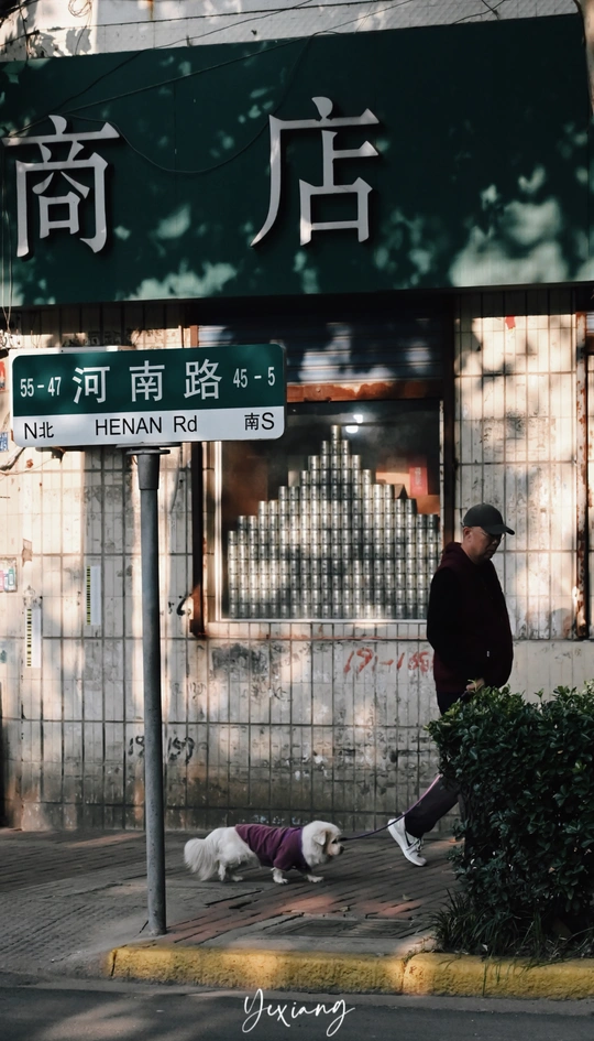 一个男子走在街上 他的狗在地上打盹 狗身前有一栋楼上的标志。