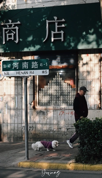 一个男子走在街上 他的狗在地上打盹 狗身前有一栋楼上的标志。