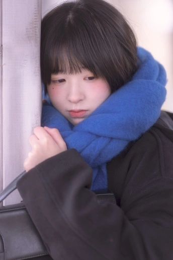 一位戴蓝围巾的年轻女子靠在窗户边 手里拿着一个包。