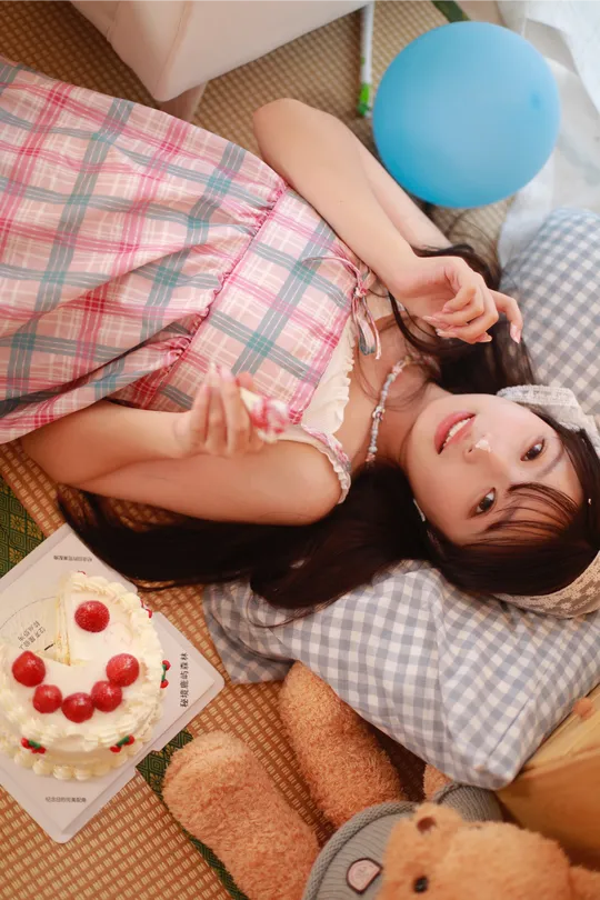 一个年轻女子躺在地板上 旁边有一个泰迪熊、一个生日蛋糕和一只蓝色气球。