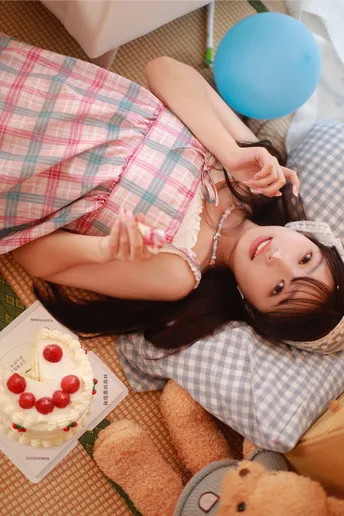 一个年轻女子躺在地板上 旁边有一个泰迪熊、一个生日蛋糕和一只蓝色气球。