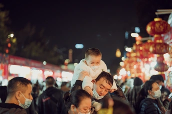 一位戴口罩的女人把婴儿背在肩膀上 晚上 一群人在繁忙的街道上走动。