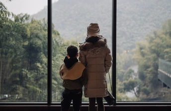 一位妇女和一个年轻男孩站在一扇大型窗户前 眺望着外面的山。