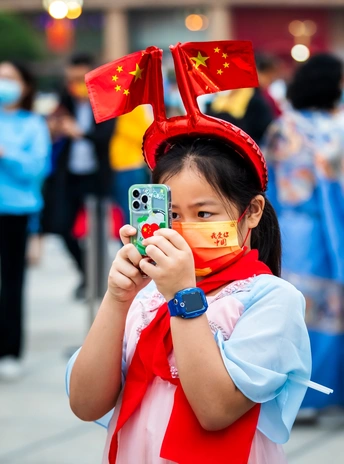 一个戴着口罩、脸上挂着中国国旗的年轻女孩拿着一部智能手机 与背景中的一个人合影