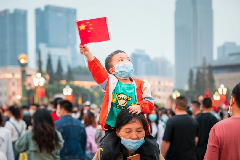 一名戴着口罩的年轻女子在街上的人群中挥舞着一面中国国旗 肩上扛着一个孩子