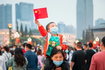 一名戴着口罩的年轻女子在街上的人群中挥舞着一面中国国旗 肩上扛着一个孩子