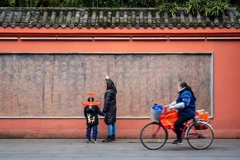 一位妇女骑着自行车 旁边站着一名儿童 还有一个人正在街上走。