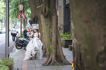 婚礼上的女士穿着婚纱走下人行道 新郎新娘站在树木旁边 摩托车已经停好。