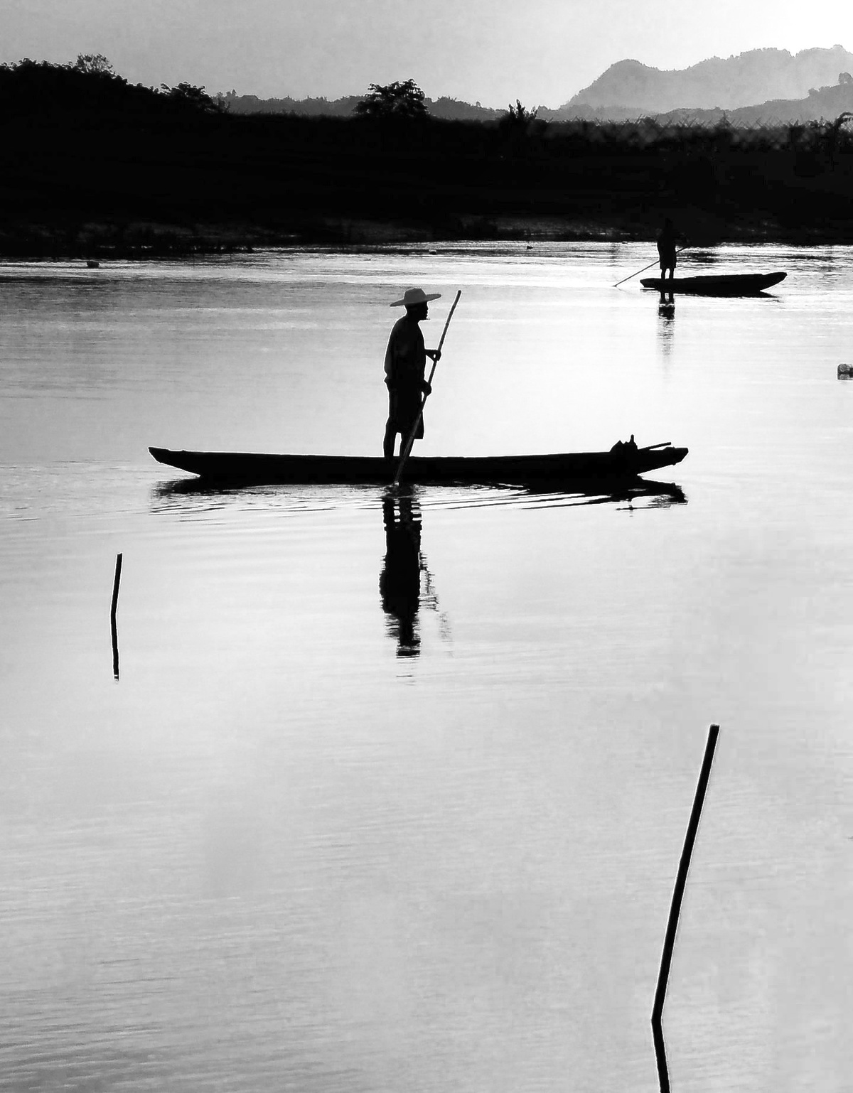 黑白照片中 一个人站在一艘小船上 身处水域 而远处另一个人在钓鱼。