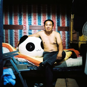 一个男人坐在床上 没有穿衬衫 房间里有一个风扇和毛绒玩具